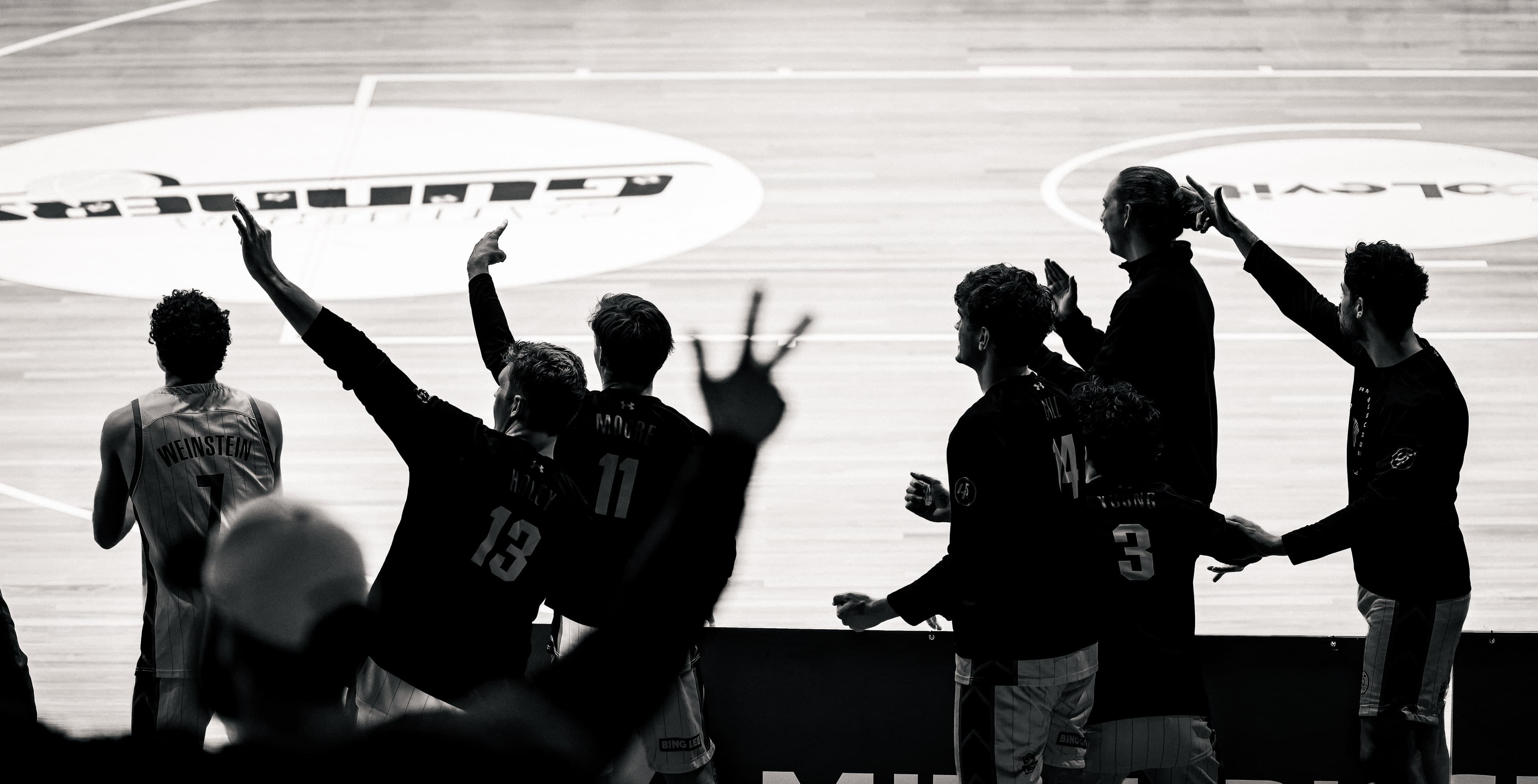 Bench players in silhouette hold up three point signs against a bright court