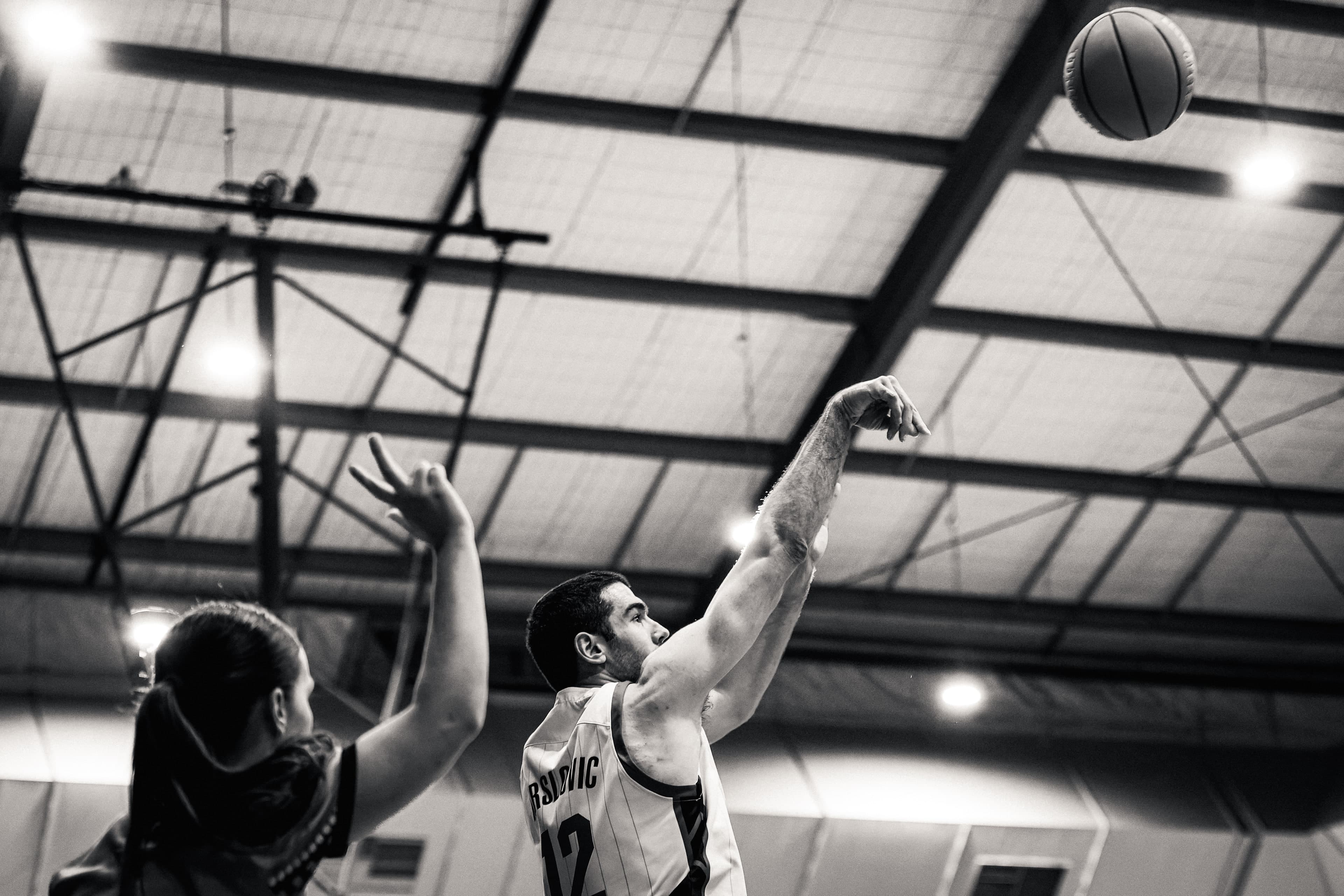 A jump shot hangs in the air beneath the roof trusses at an NBL1 East game in Canberra, in black and white