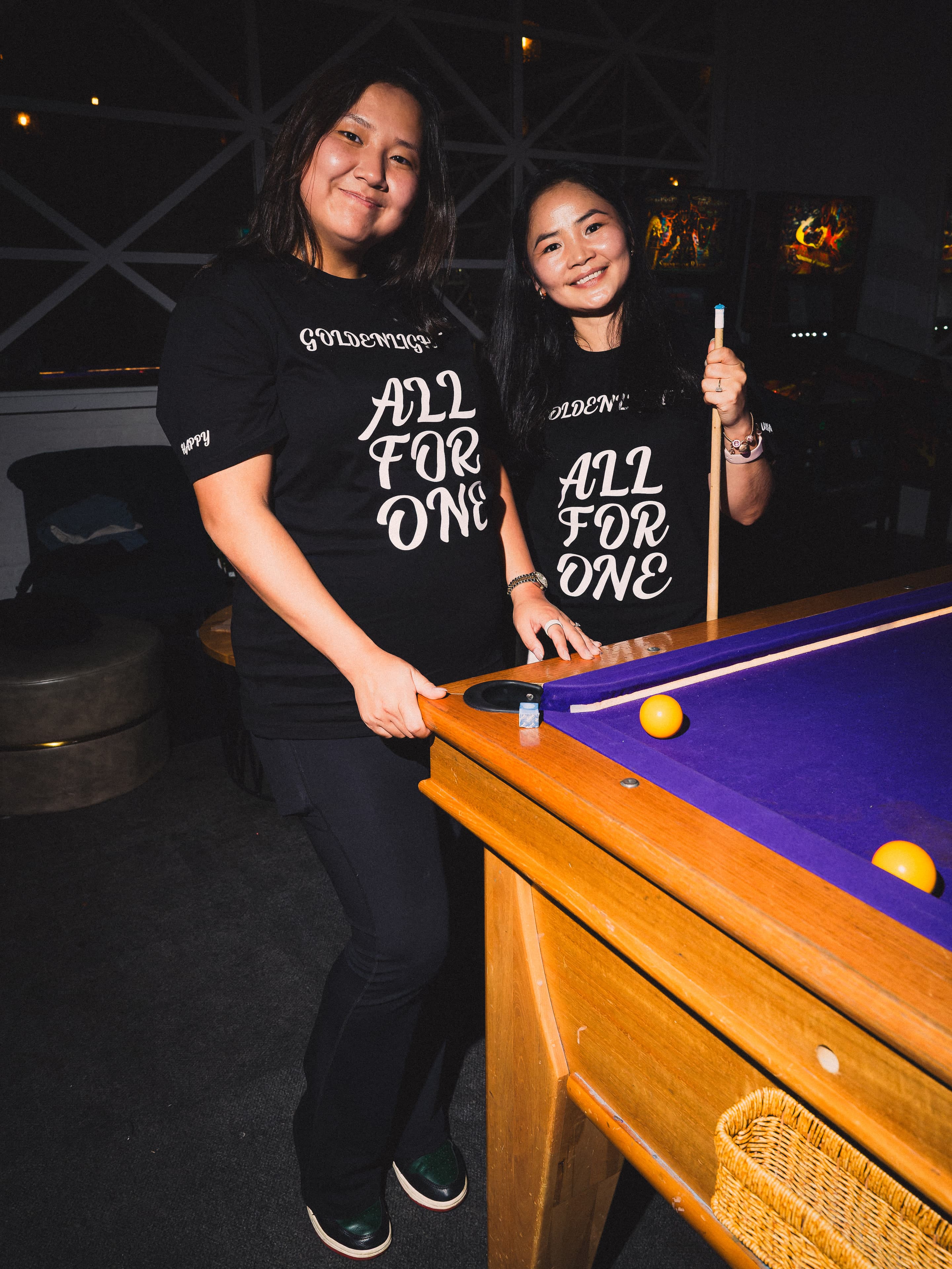 Two women in All For One tees at the pool table, one holding a cue upright
