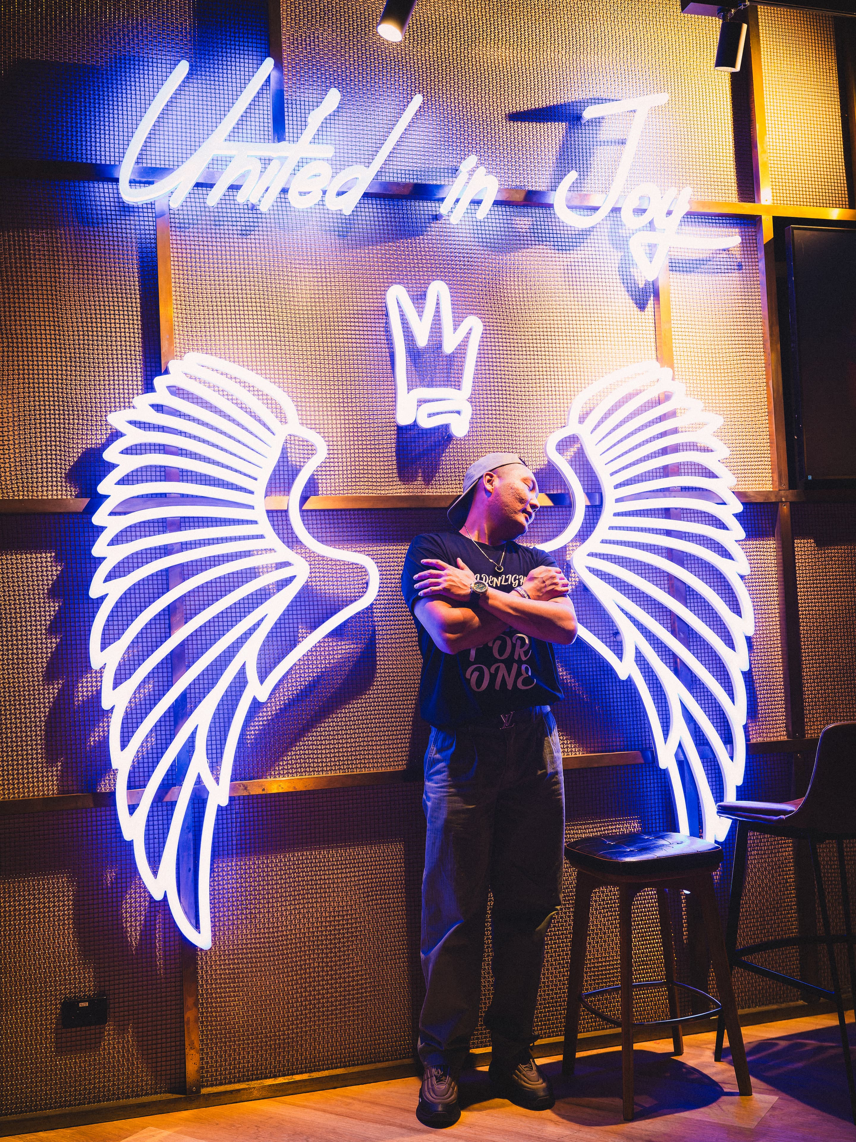 A man stands between the neon wings with his arms crossed, United in Joy lit blue above him