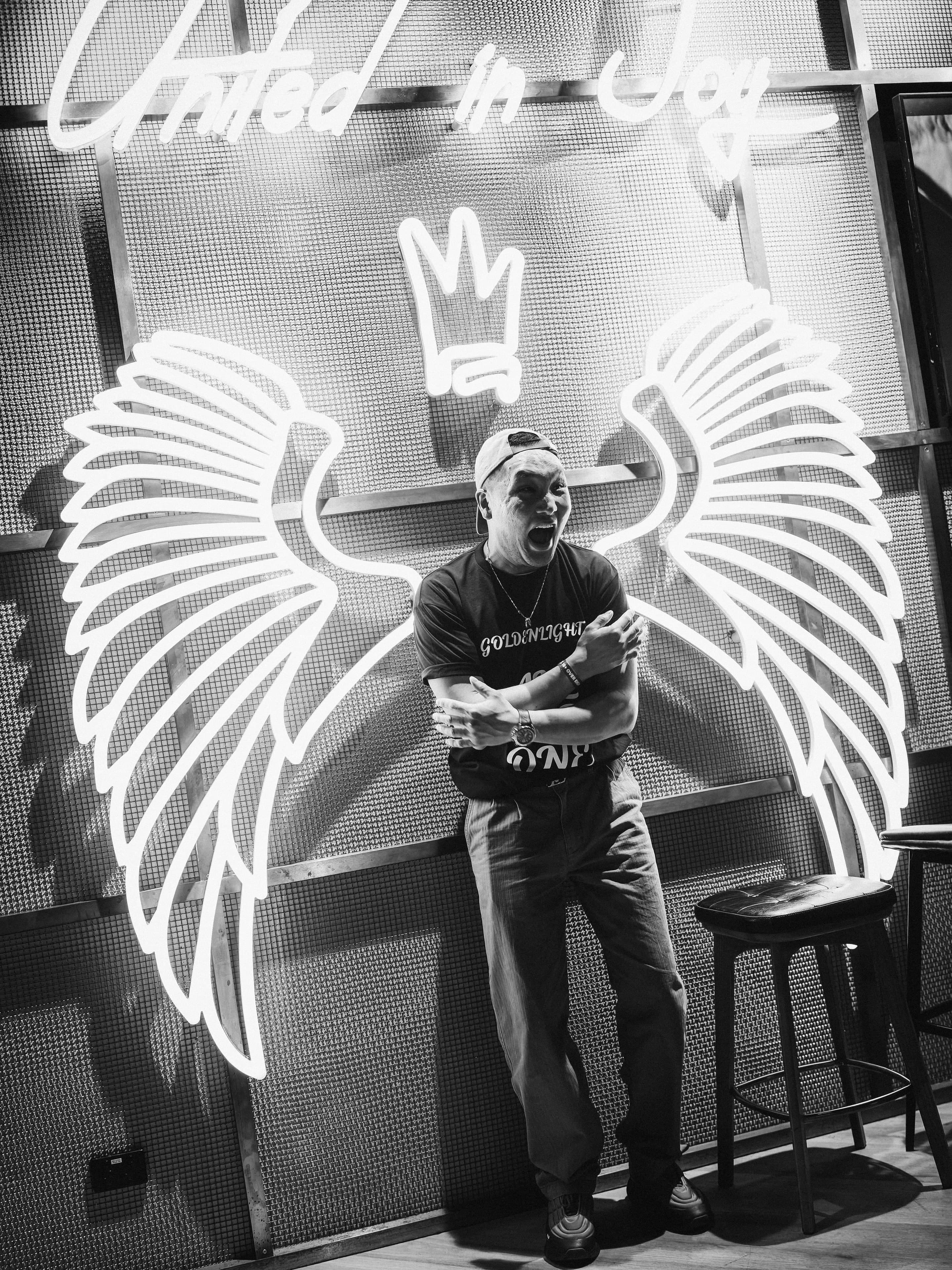 A man in a backwards cap shouts under the neon wings, United in Joy lit above him, in black and white