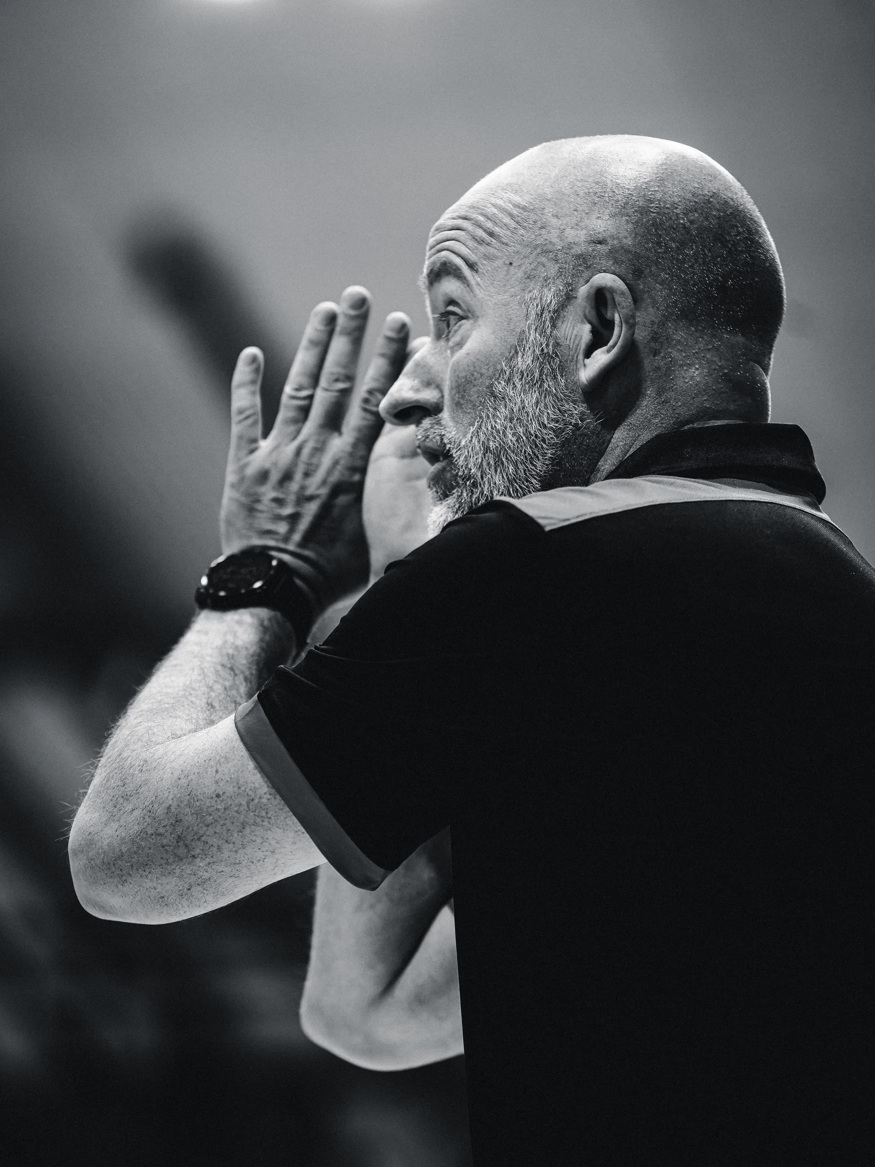 A coach cups his hands and shouts a call across the court, in black and white