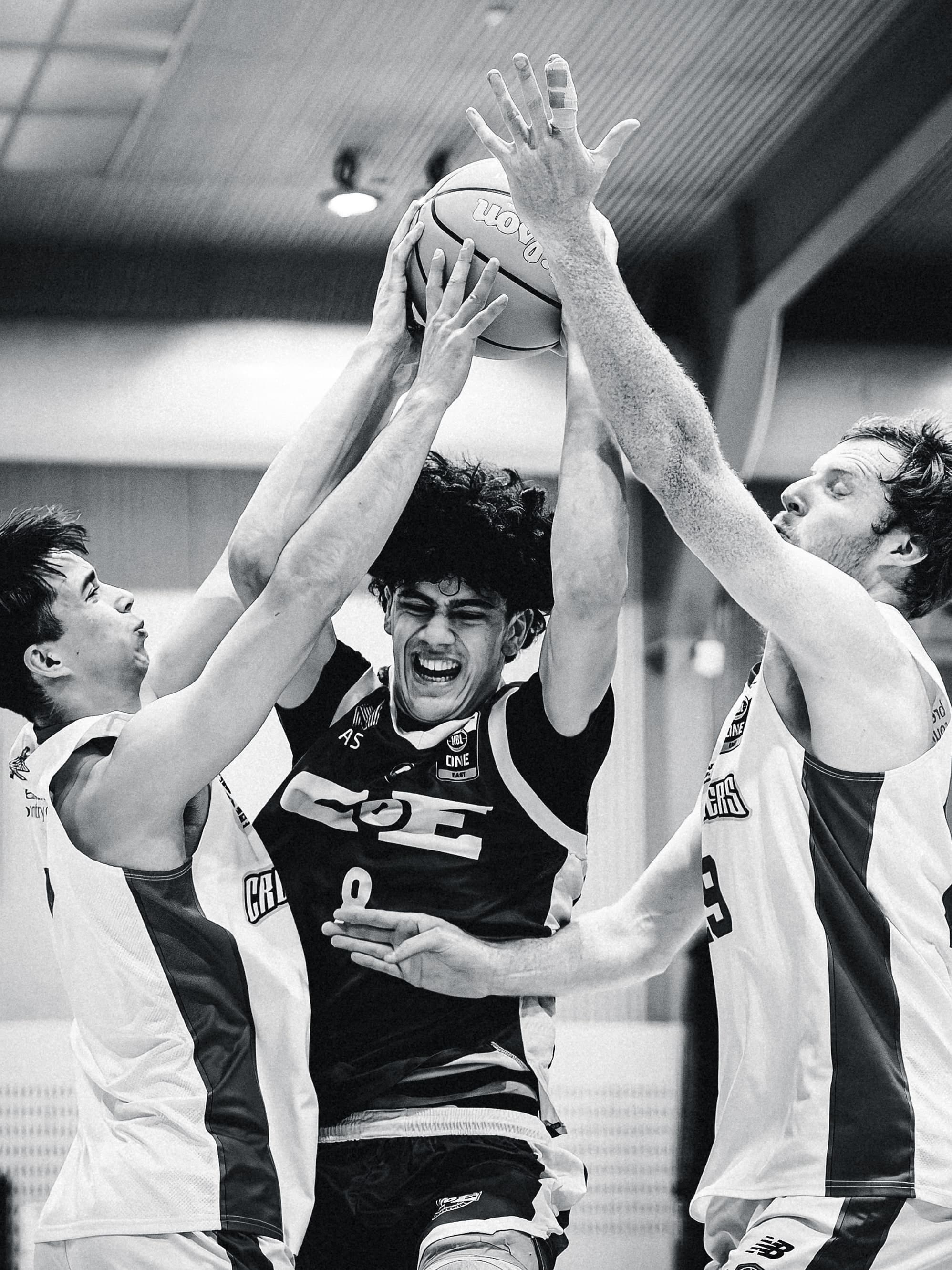 A Centre of Excellence player holds the ball above his head while two Central Coast Crusaders claw at it, in black and white