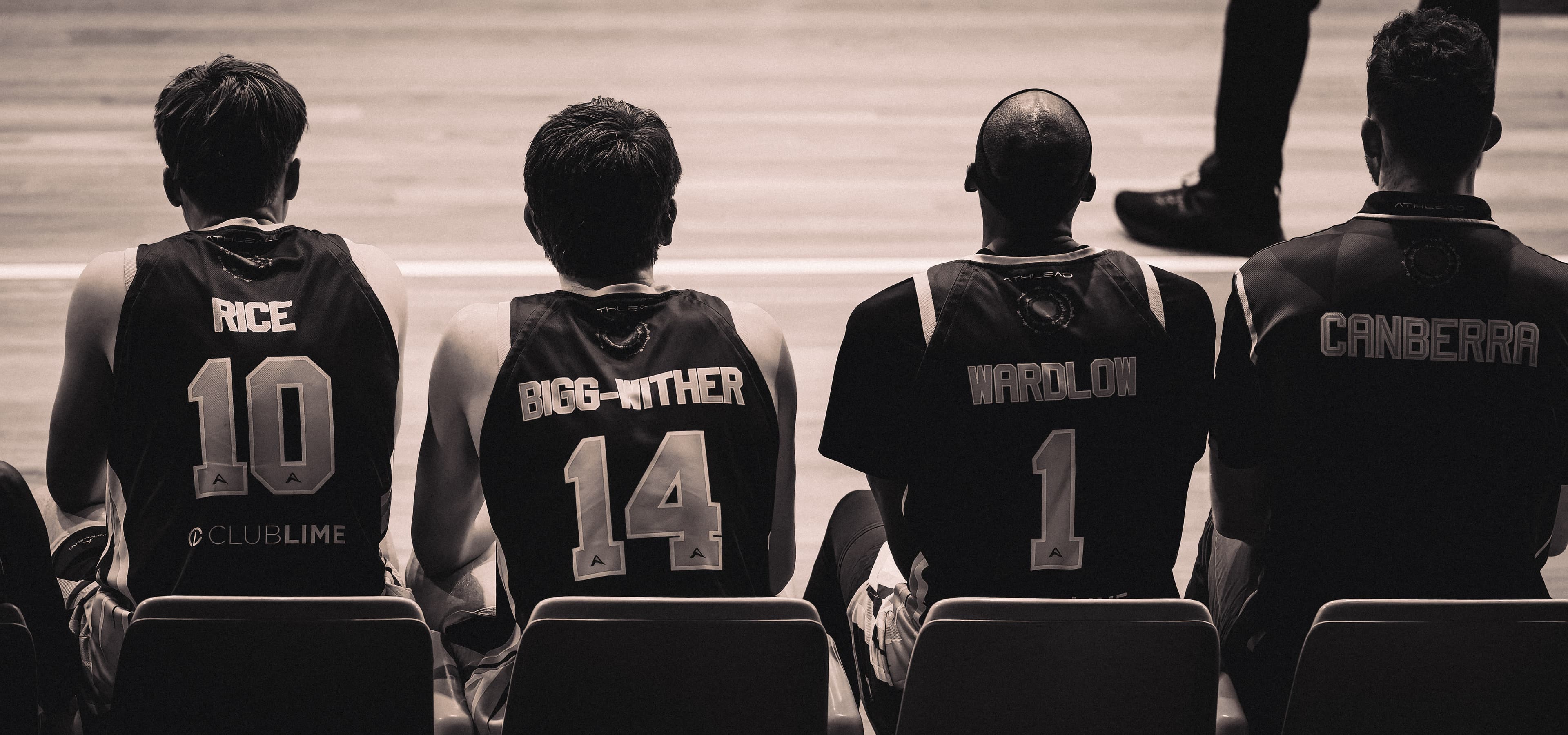 Four Canberra Gunners players sit along the bench with their names across their backs, in black and white