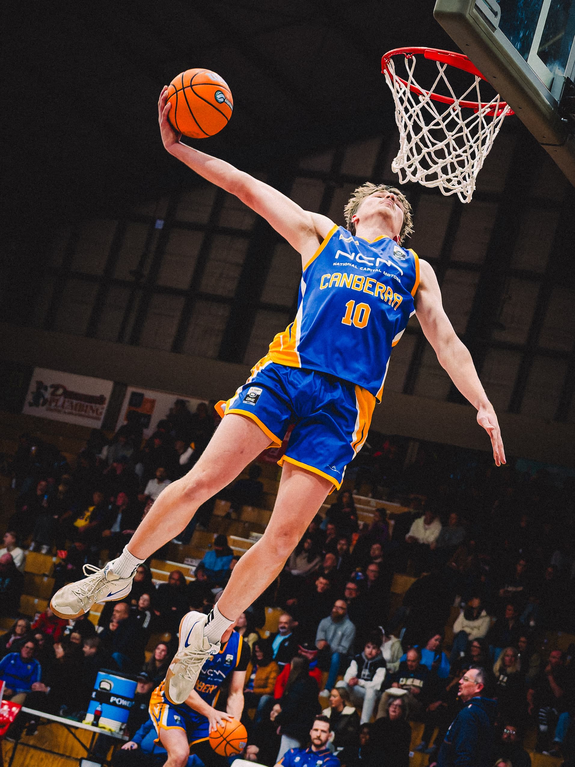 A Canberra Gunners guard climbs to the ring with the ball held back behind his head, crowd below him