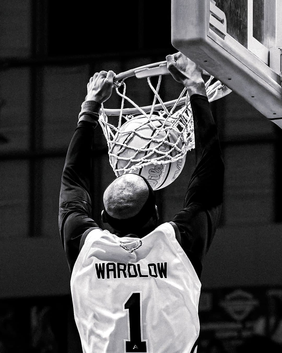 Wardlow hangs on the rim after a dunk, photographed from behind in black and white