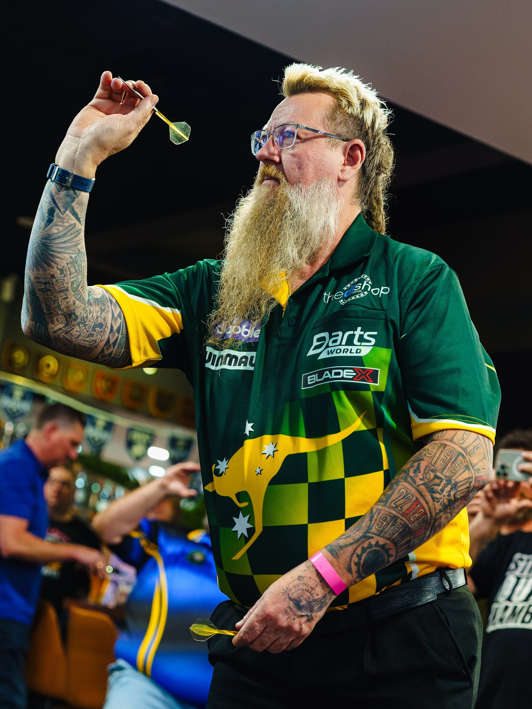A darts player in a green and gold shirt aims under the lights