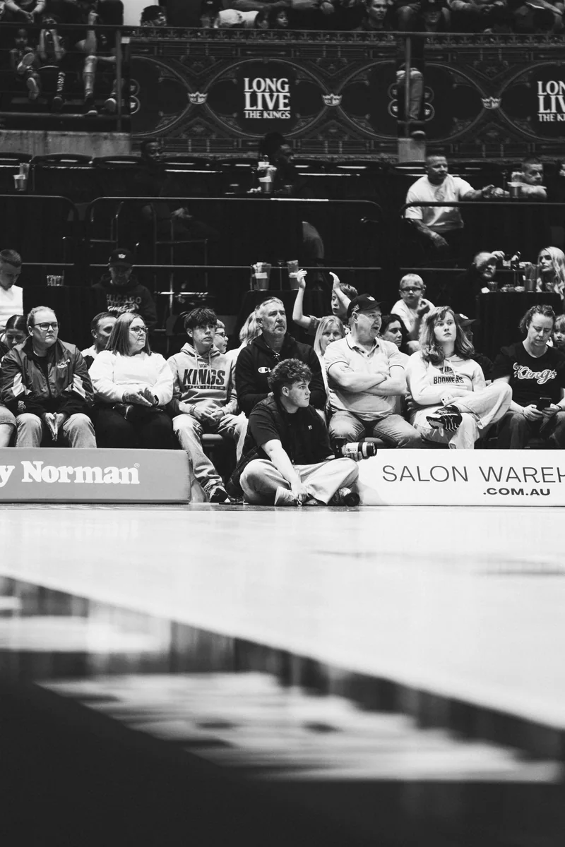 Black and white photograph of Kian sitting courtside with his camera, with spectators behind him