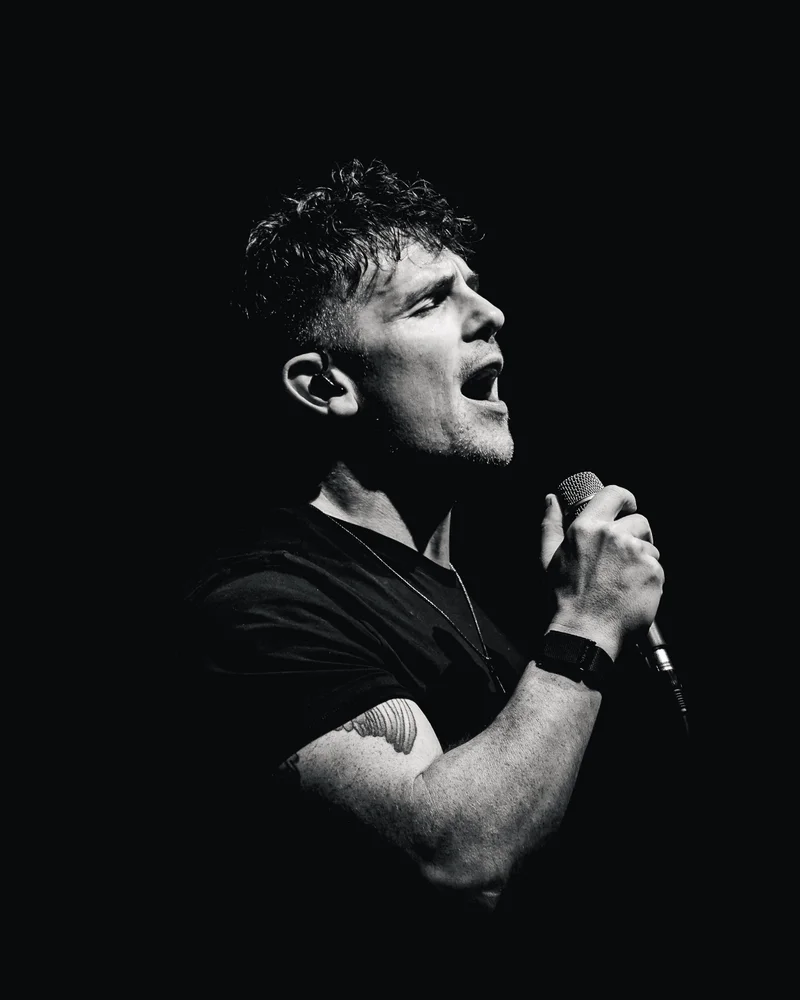 Michael Paynter singing into a handheld microphone under stage lighting, in black and white